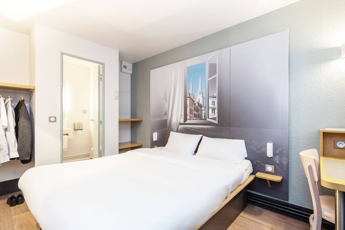 Een hotelkamer met een bed, een bureau, een stoel, een kledingkast en een groot raam met uitzicht op de stad.