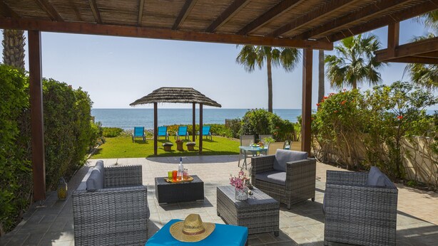 A covered outdoor seating area with wicker furniture, a table with drinks, and a view of the sea. - Beachfront Villa Meneou 5 (Larnaca)