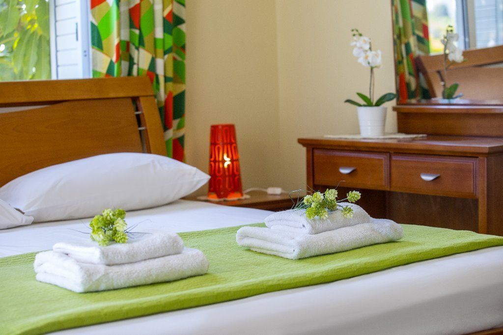 Ein ordentlich gemachtes Bett mit weißen Handtüchern und einer grünen Decke, ein Nachttisch aus Holz mit einer roten Lampe, eine Holzkommode und eine Topf-Orchidee.