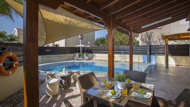 An outdoor pool area with a covered patio, wicker furniture, and a table set for dining. - Ayia Napa Tropical Beach Villa (Ayia Napa)