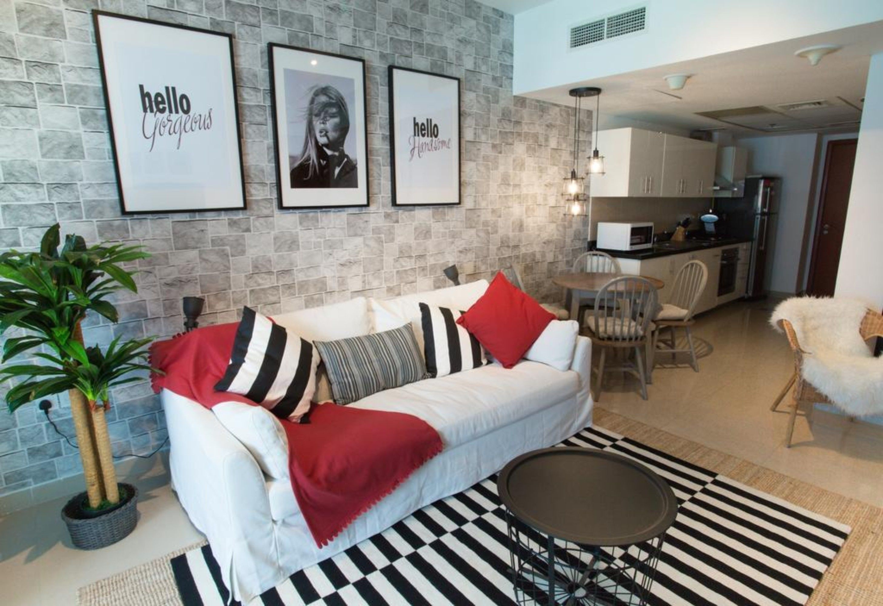 Un salon moderne avec un canapé blanc, des coussins à rayures, un plaid rouge et un tapis noir et blanc. Des tableaux encadrés ornent le mur, et une plante en pot est posée dans un coin.