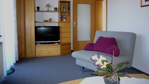 Een woonkamer met een televisie, houten meubels, een grijze bank, een paars plaid en een potorkidee.