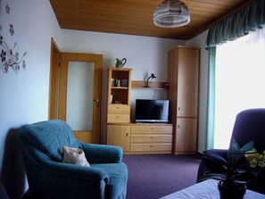 A room with a blue armchair, a television on a wooden cabinet, and a window with curtains.