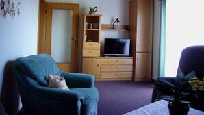A room with a blue armchair, a television on a wooden cabinet, and a window with curtains.