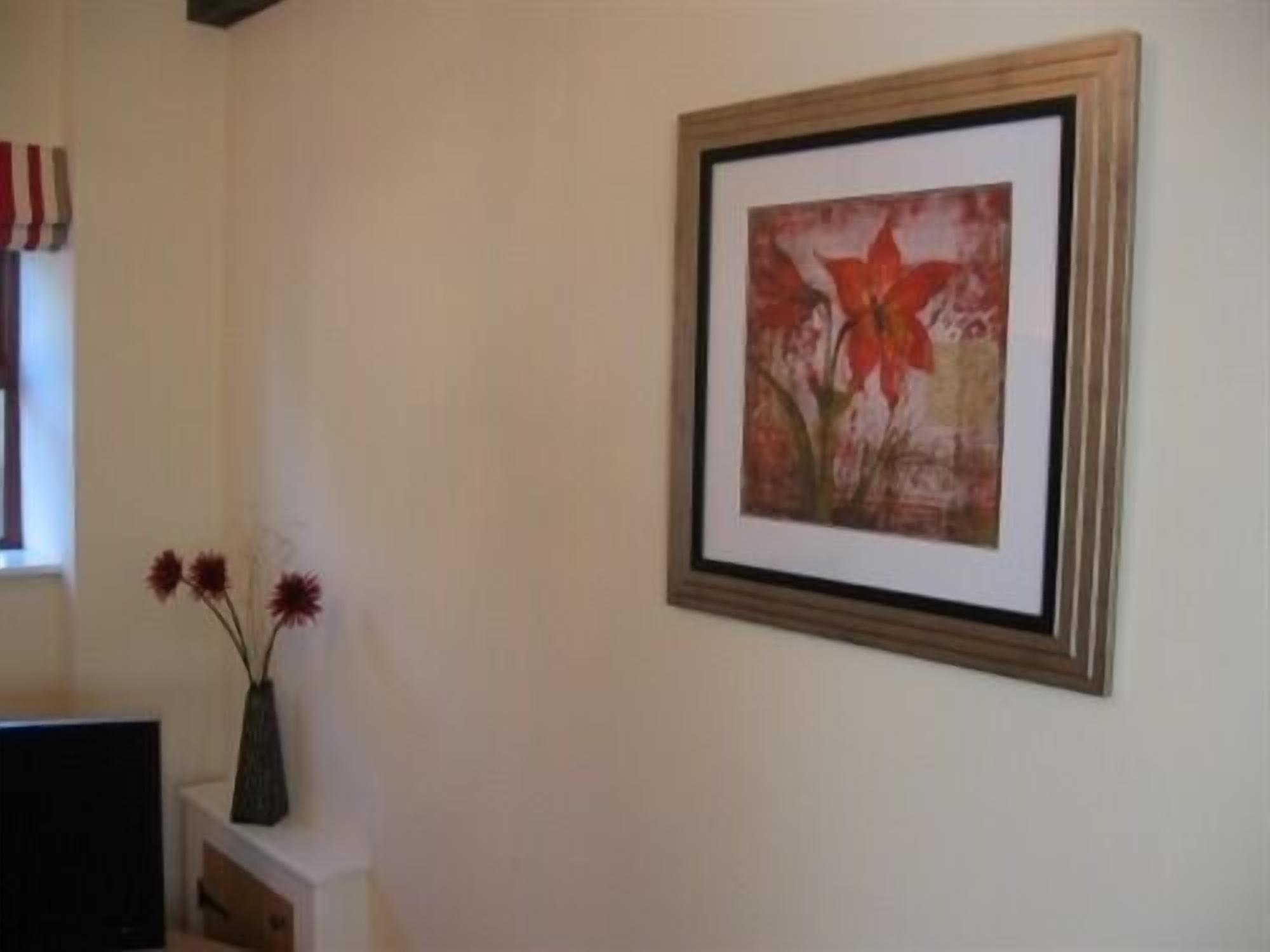 A framed painting of flowers on a wall, a vase with flowers on a white console table, and a television on a stand.