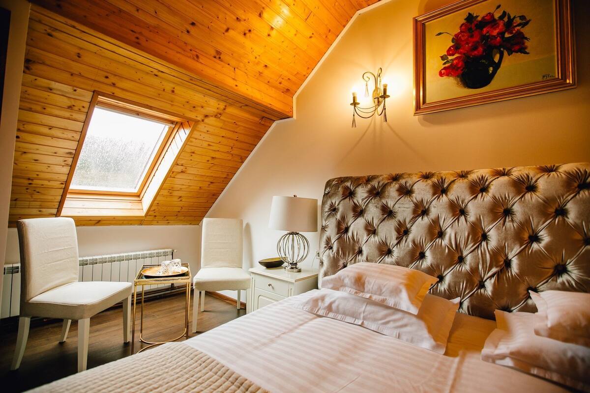 Ein gemütliches Schlafzimmer mit schrägen Wänden, einem großen Bett mit einem gepolsterten Kopfteil, einem Nachttisch mit Lampe, einem Sessel und einem Fenster, das für natürliches Licht sorgt.