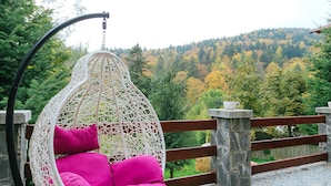 A white wicker hanging chair with pink cushions on a tiled patio overlooking a forested hillside. - Casa Natalia (Sinaia)