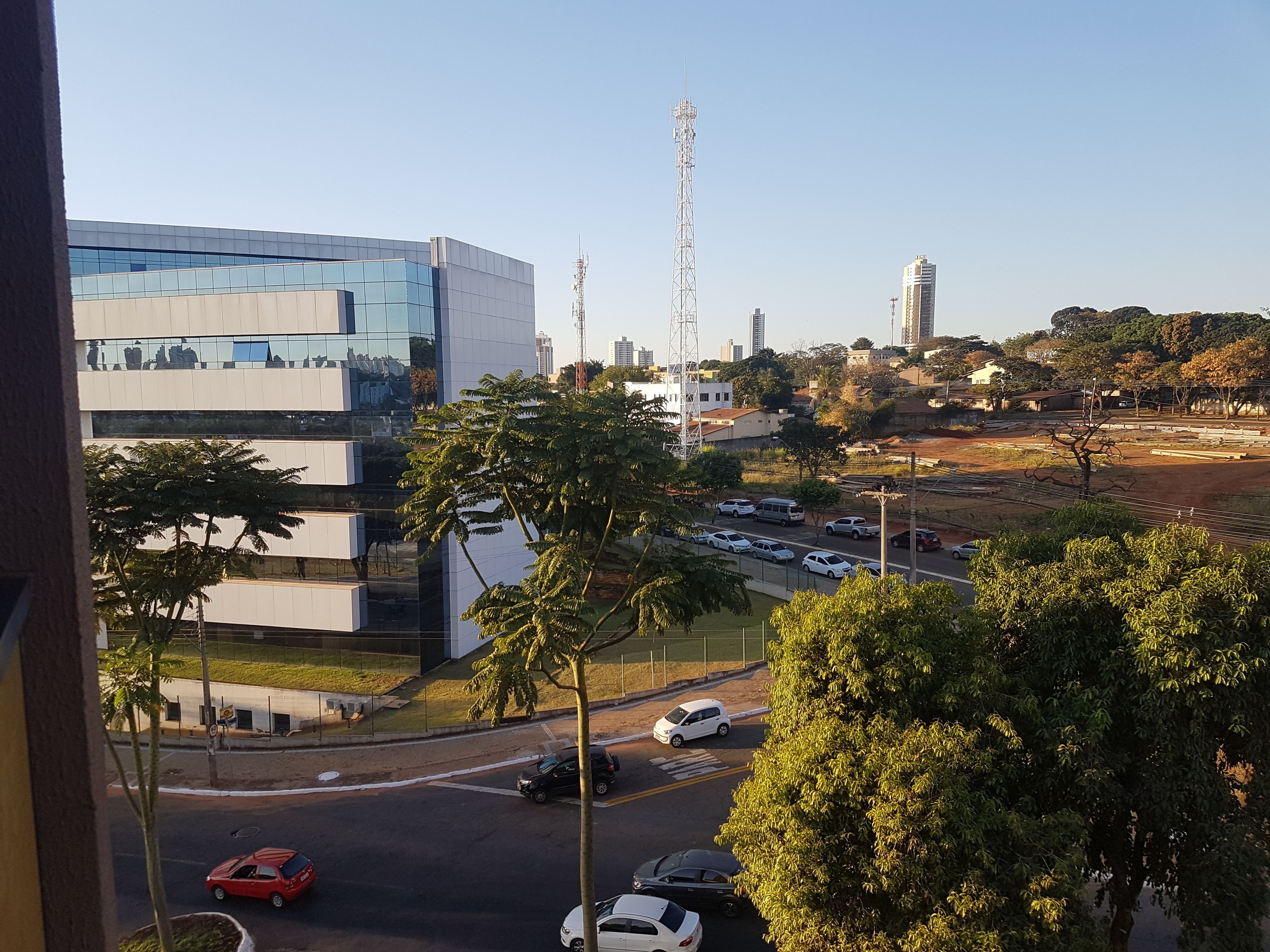Um edifício moderno com fachada de vidro, um estacionamento com vários carros e uma vista clara da paisagem urbana ao redor.