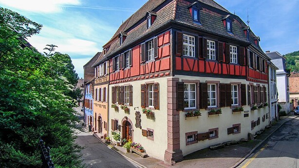 Un edificio tradizionale a struttura in legno e intonaco, con tetto spiovente, numerose finestre con persiane e un piccolo portico con fioriere.