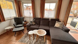 A living room with a grey sofa, a grey armchair, a small white table, and a coffee table. There are curtains on the windows, and a lamp is on a side table. - Bright, cozy apartment, centrally located in a quiet area of Zingst with WLAN (Mecklenburg-Vorpommern)