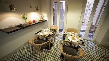 Uma sala de jantar com chão quadriculado, cadeiras de vime, mesa de madeira posta para dois e prateleira com utensílios de cozinha.