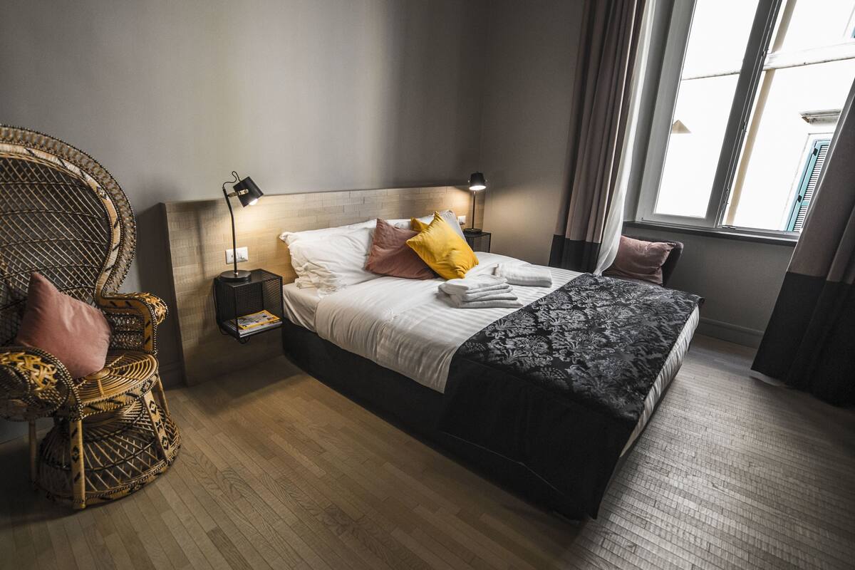 Ein Schlafzimmer mit einem großen Bett, einem Korbstuhl, einem Nachttisch und einem Fenster mit Vorhängen.