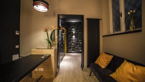 Un interior moderno con un escritorio de madera, un sofá negro con cojines amarillos, una planta y una puerta que da a una habitación con papel tapiz floral blanco y negro.