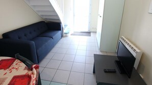 A room with a blue sofa, a small table with a TV and remote, a staircase, and a door leading outside. - Cottage near the sea with parking and pets allowed (Siouville-Hague)
