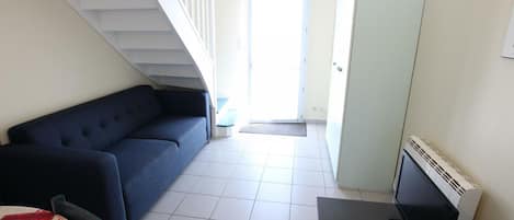 A room with a blue sofa, a small table with a TV and remote, a staircase, and a door leading outside.