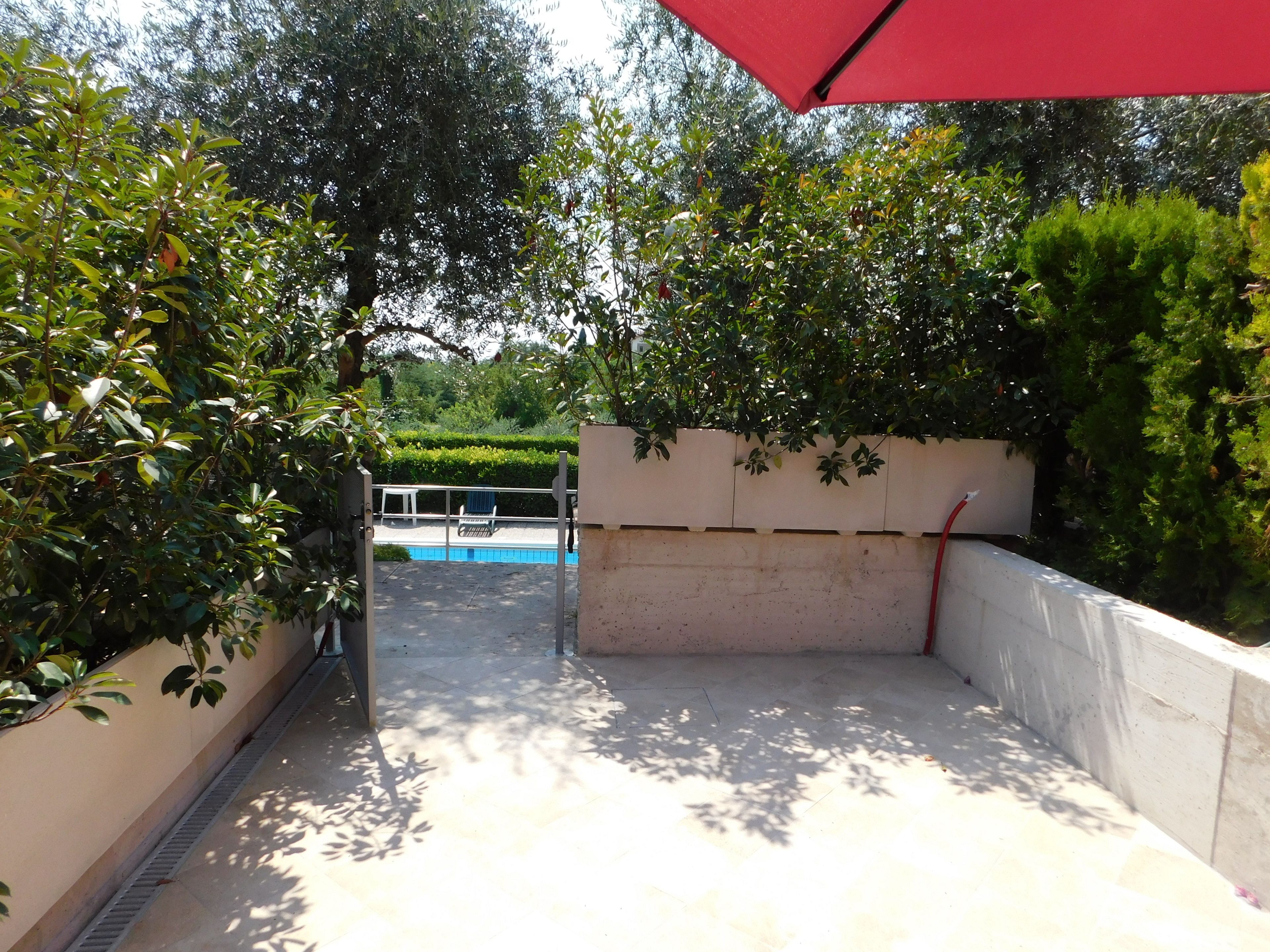 Un área de piscina con un muro de concreto, una sombrilla roja y follaje circundante.