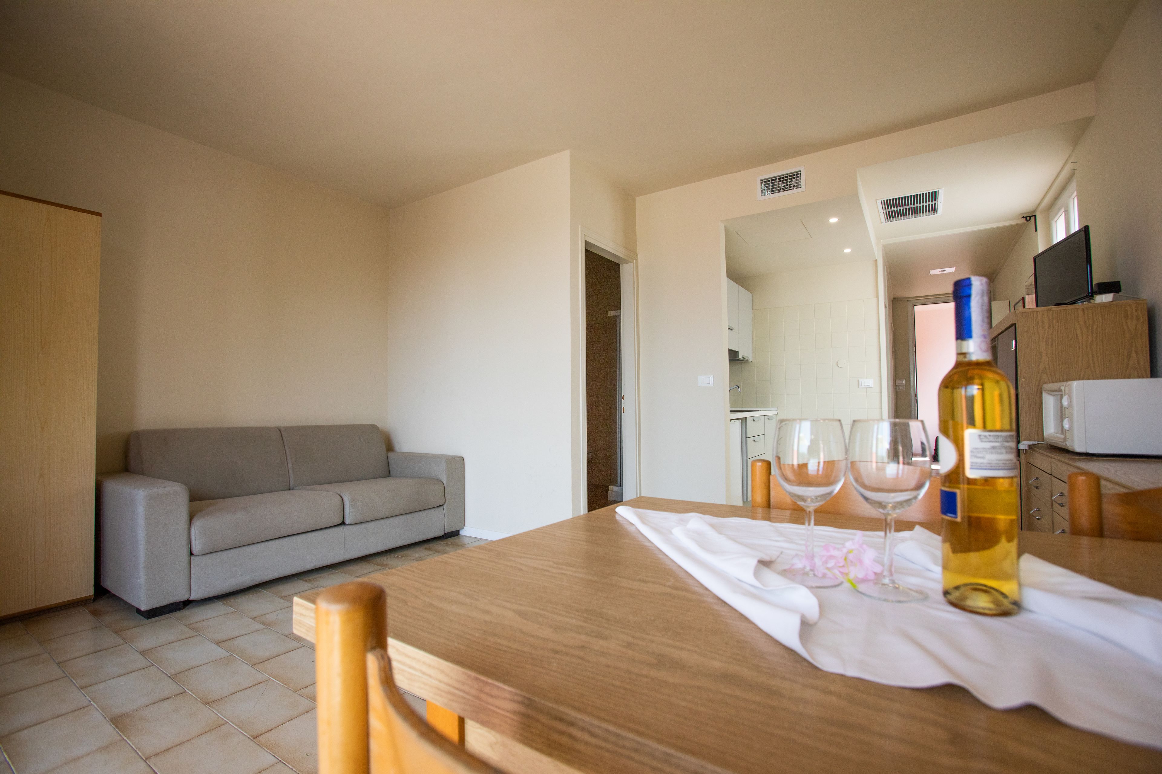 Ein modernes Wohnzimmer mit einem Holzesstisch, zwei Weingläsern und einer Weinflasche. Ein graues Sofa steht an der Wand, und im Hintergrund ist eine Küchenzeile erkennbar.
