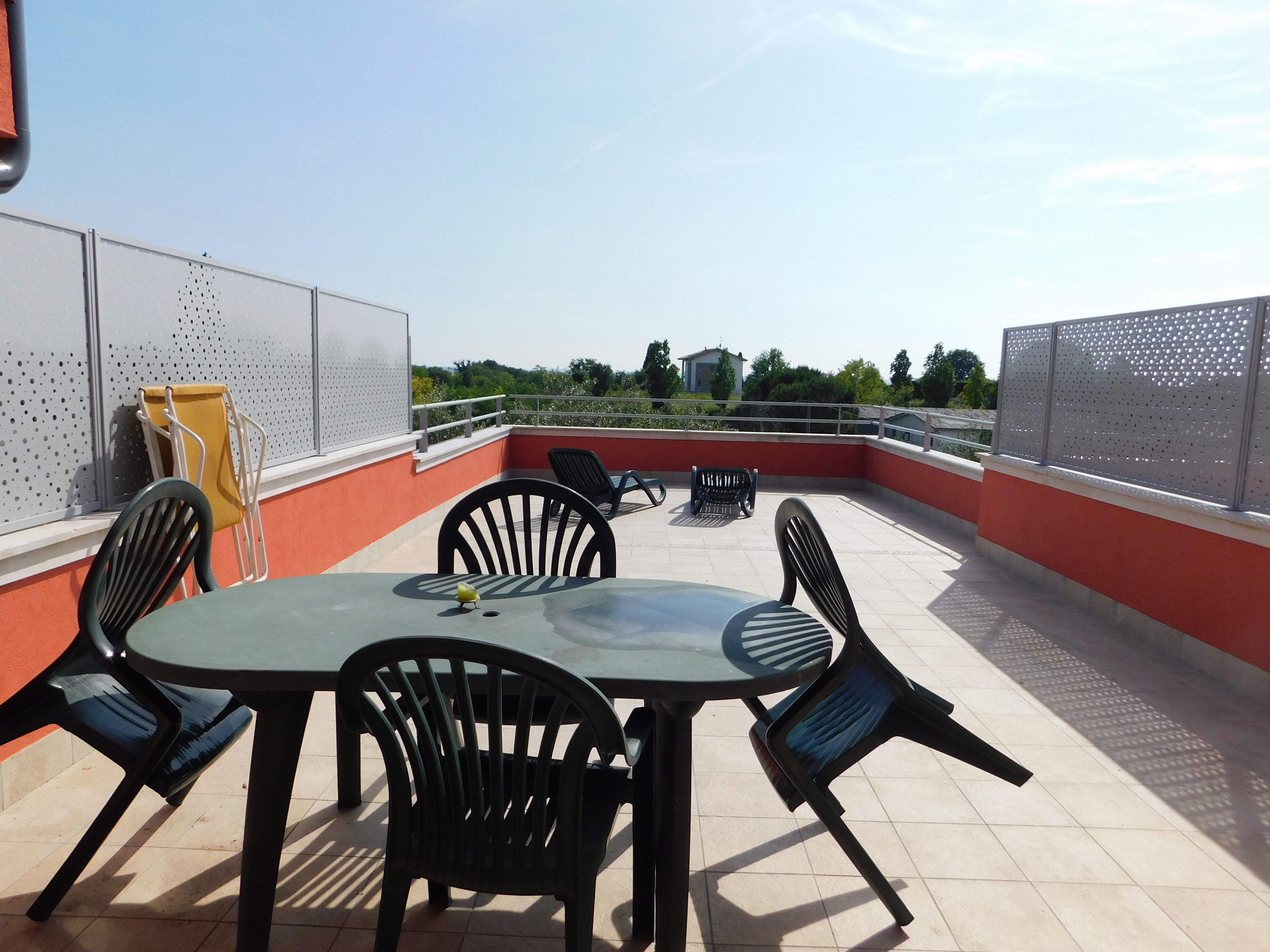 A rooftop terrace with a table and chairs, a yellow chair, and a view of greenery and buildings in the distance.