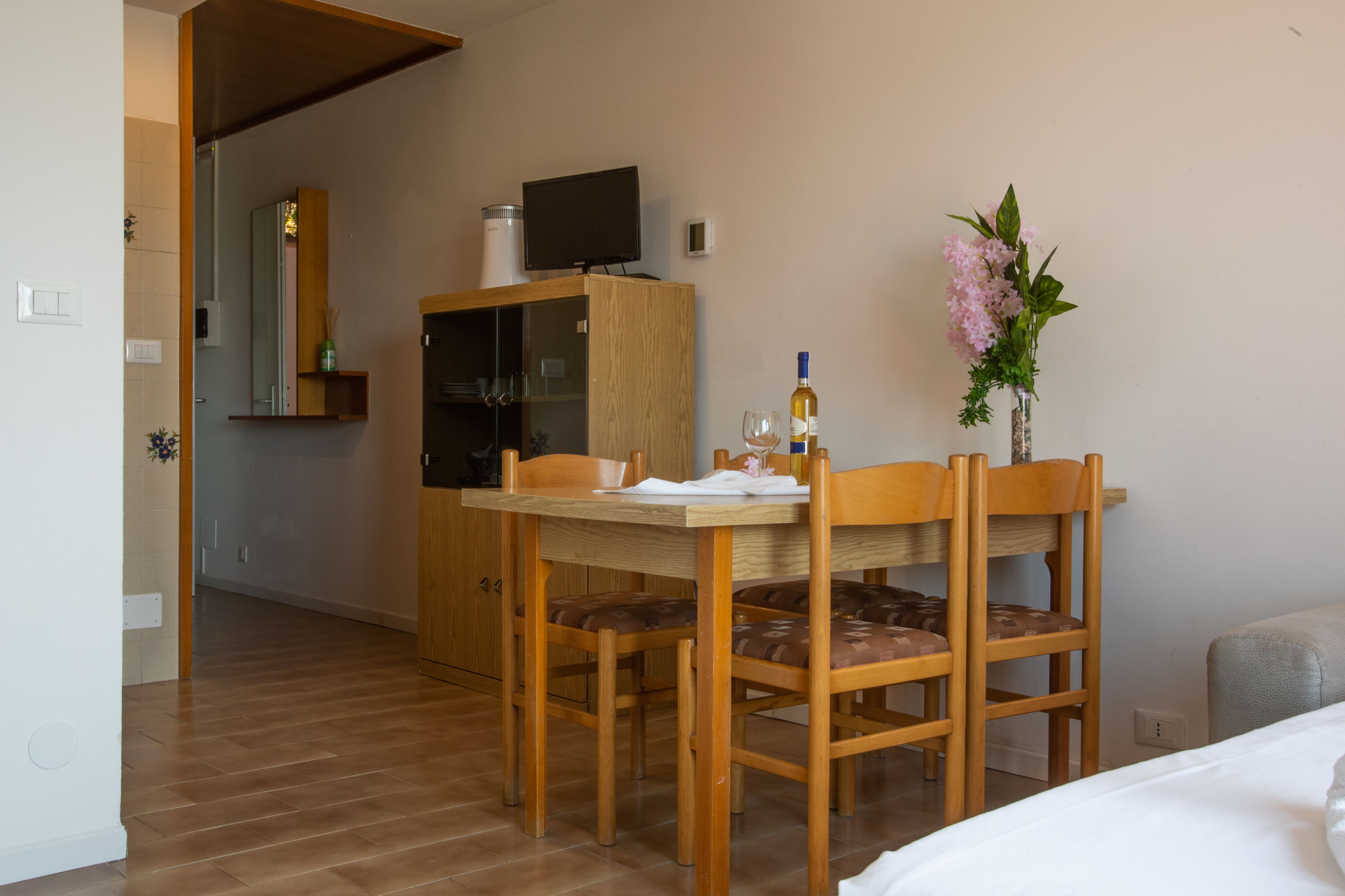 A hotel room with a wooden dining table, chairs, a TV on a cabinet, a vase with flowers, and a bed.