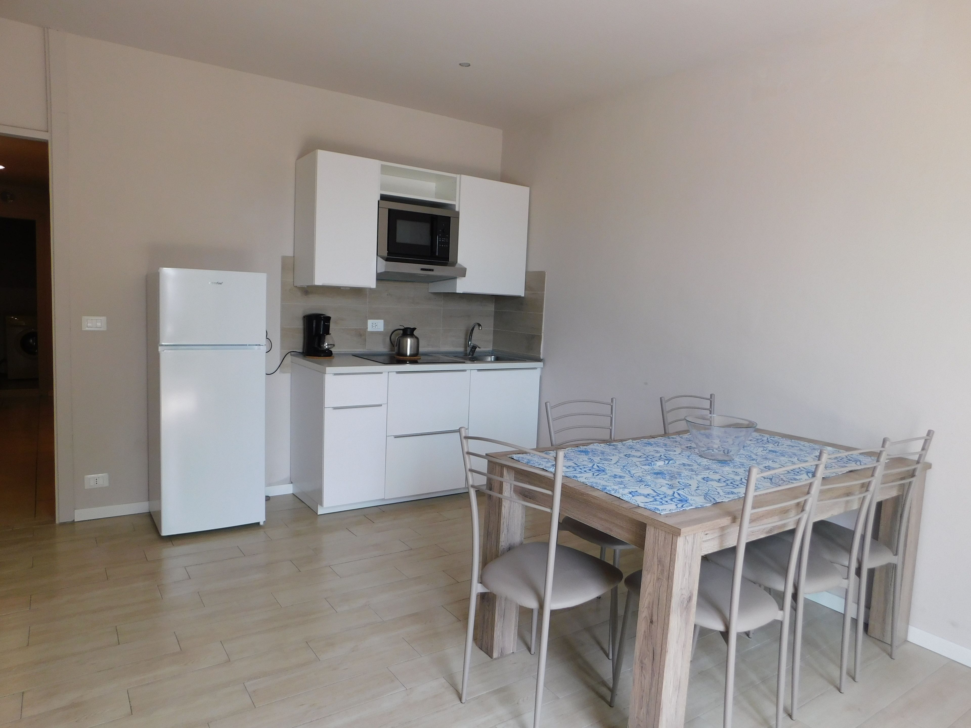 Een kleine keuken met witte apparaten, een eettafel met stoelen en een tafelkleed met dessin.