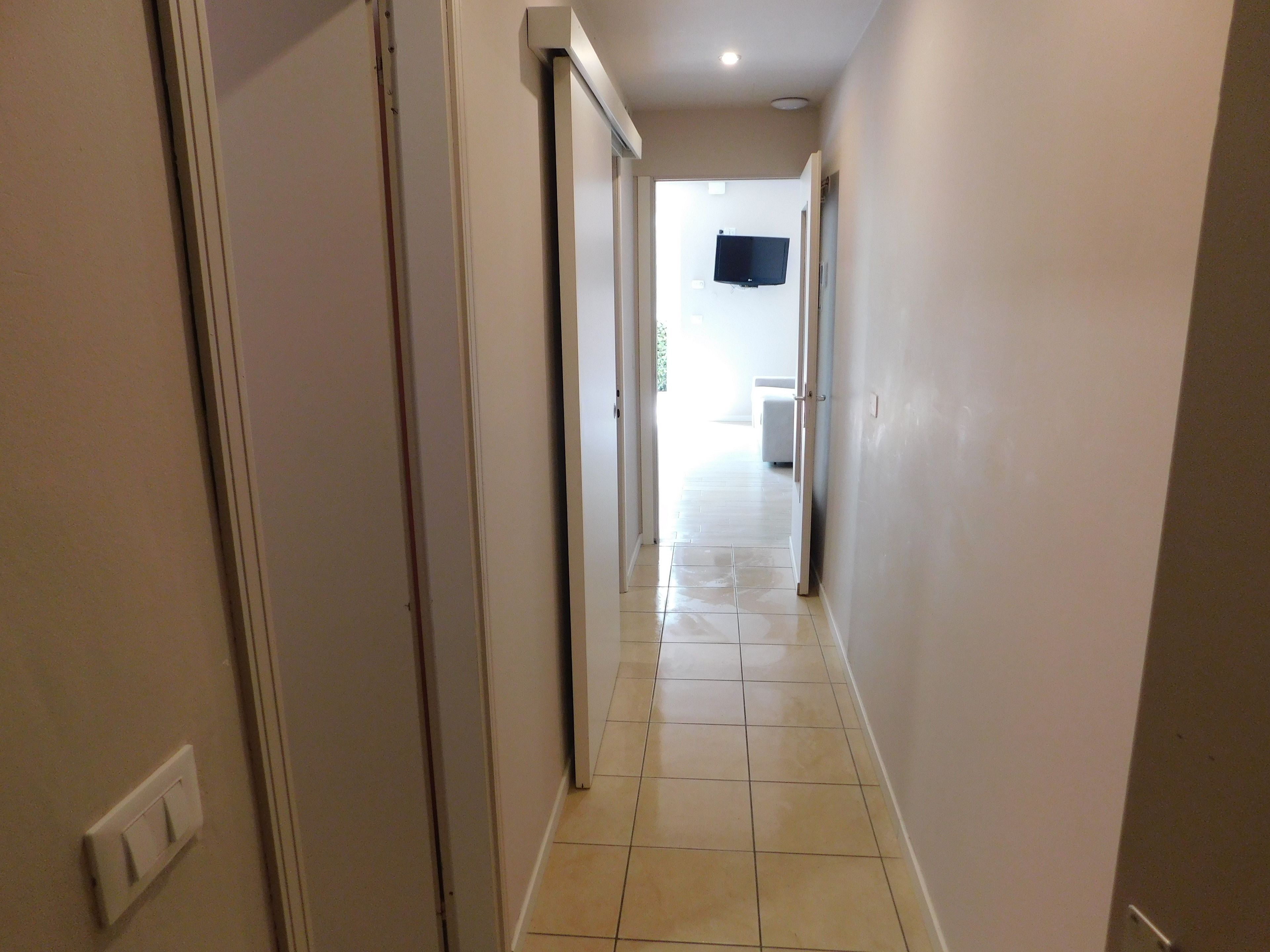 A narrow hallway with a tiled floor, leading to a room with a television mounted on the wall.