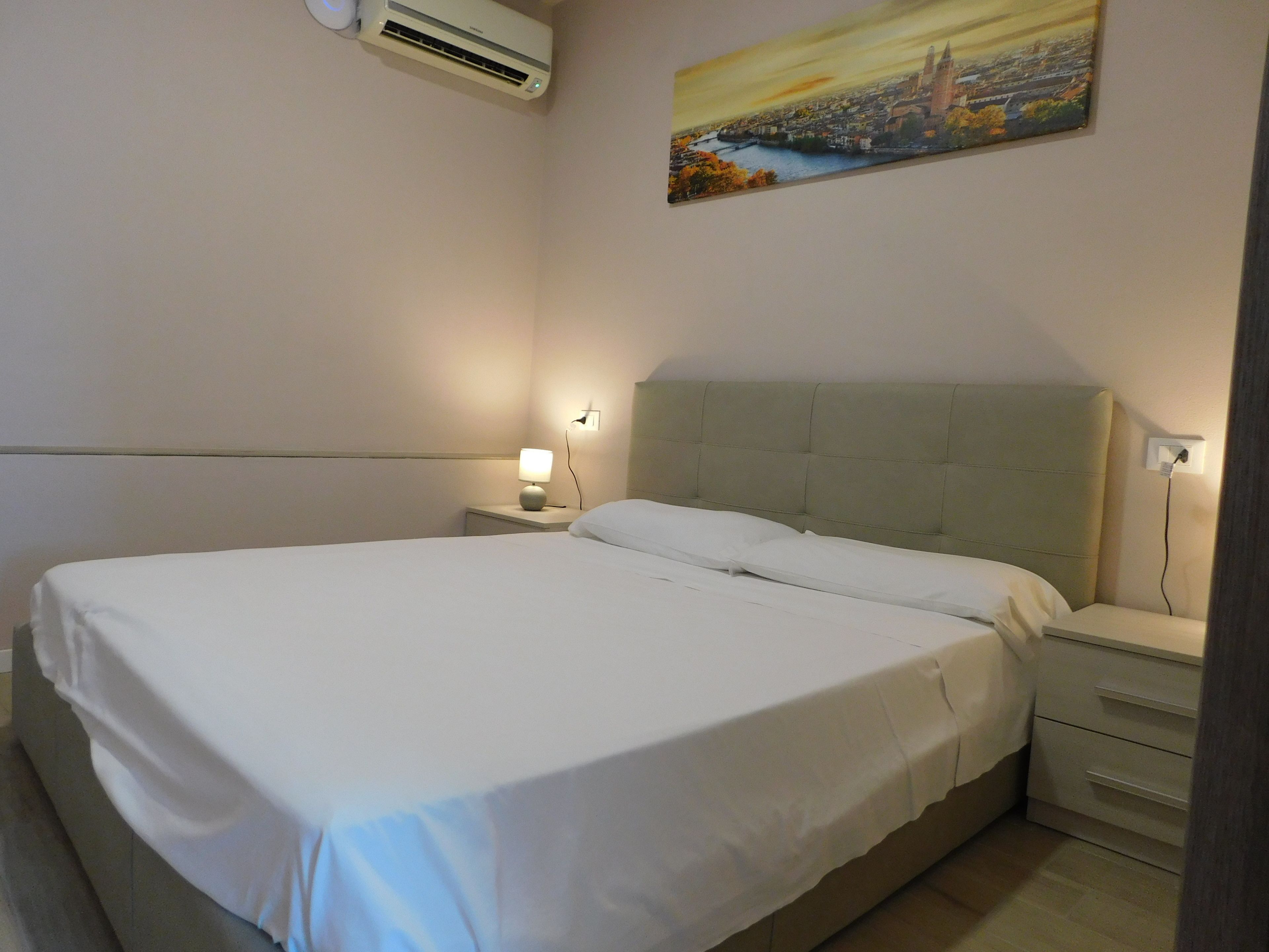 Un letto rifatto con cura in una camera d'hotel, con un quadro raffigurante un panorama urbano sulla parete, una lampada da comodino e un condizionatore sopra il letto.