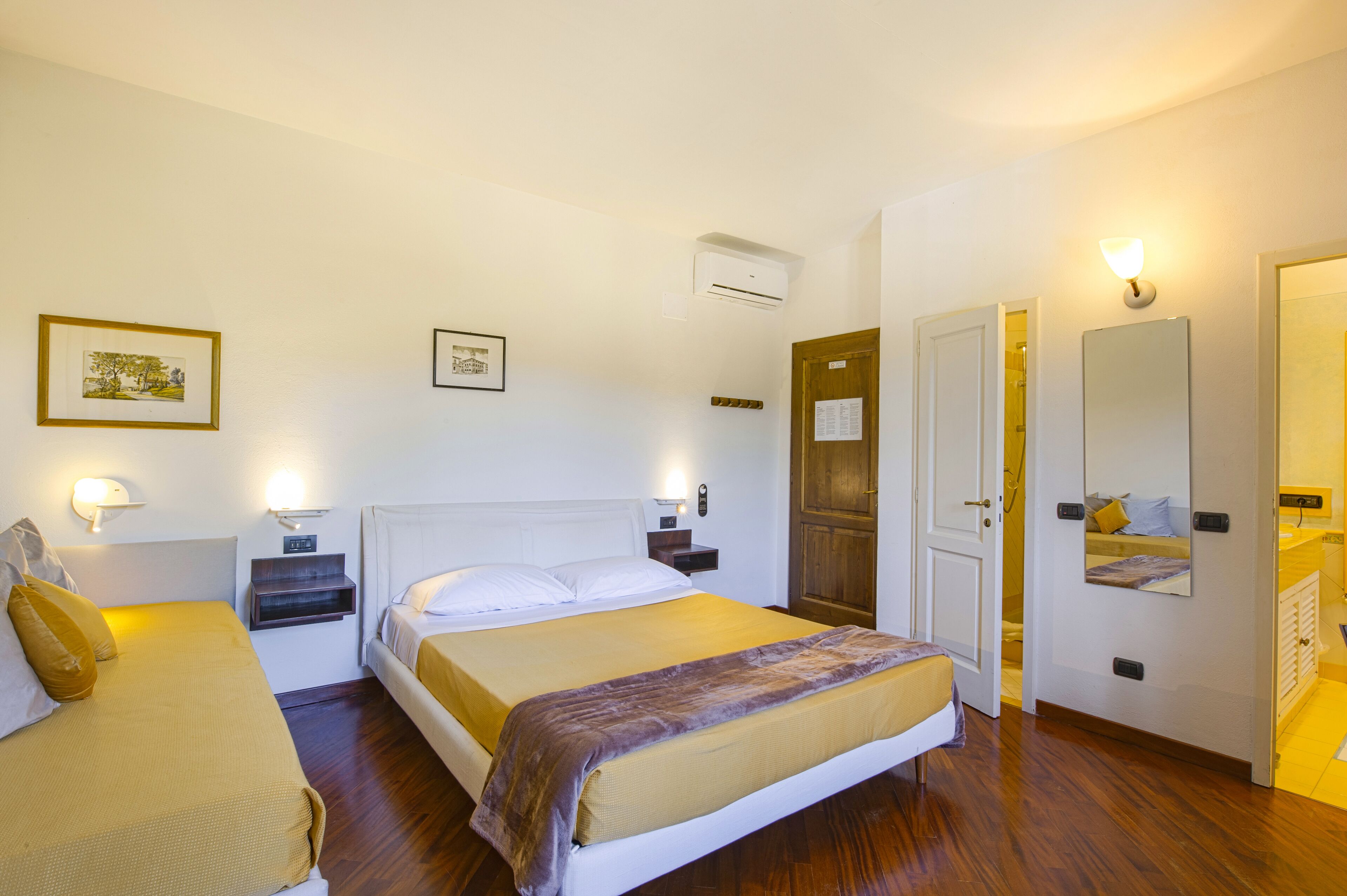 A hotel room with two beds, a wooden floor, a mirror, and a door leading to a bathroom.