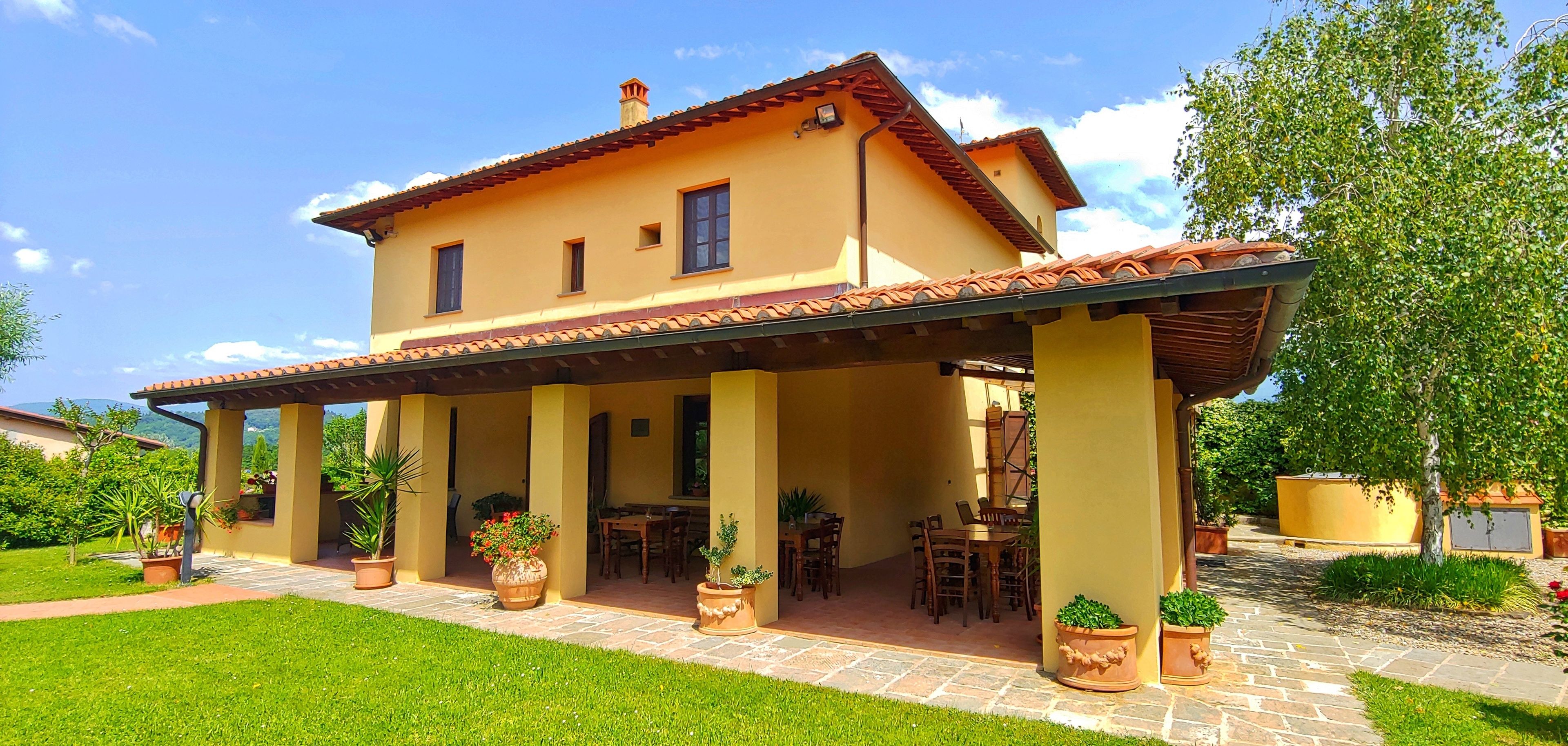 Ein zweistöckiges Haus mit überdachter Terrasse, Sitzgelegenheiten im Freien und Pflanzgefäßen.