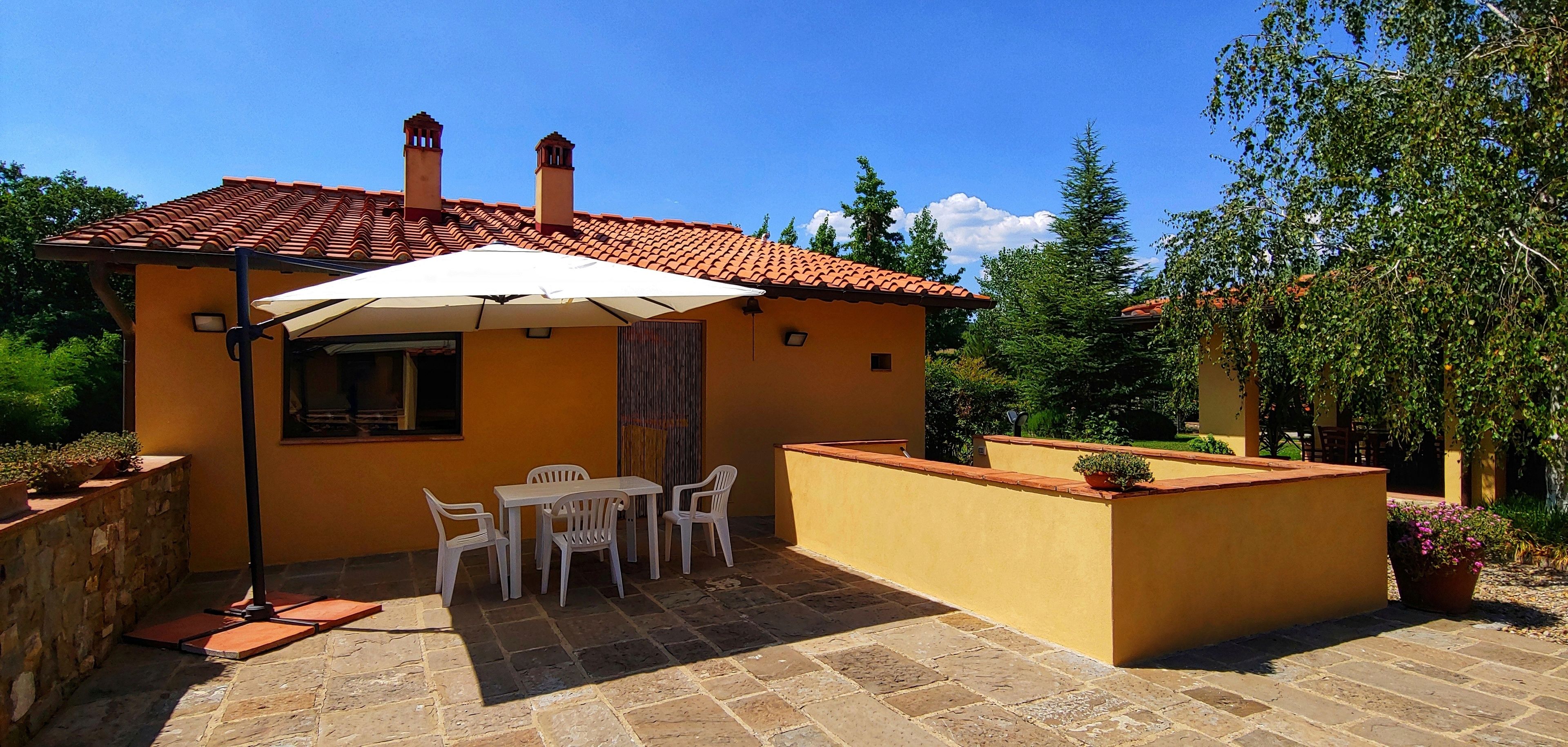 A small yellow building with a tiled roof, a patio with a table and chairs, and a large umbrella.