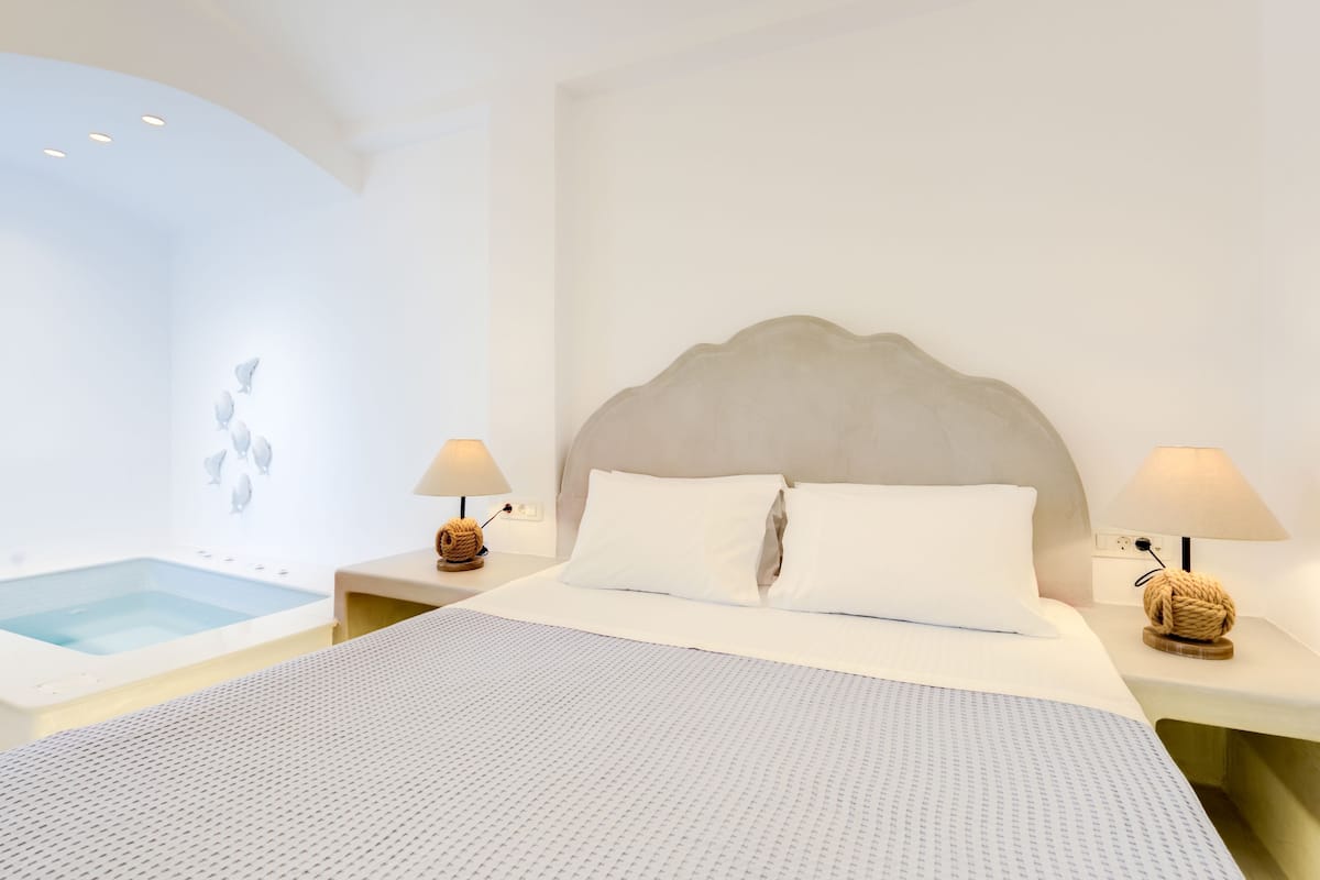 Una habitación de hotel moderna con una cama grande, dos mesitas de noche con lámparas, un cabecero empotrado y una pequeña zona de piscina.