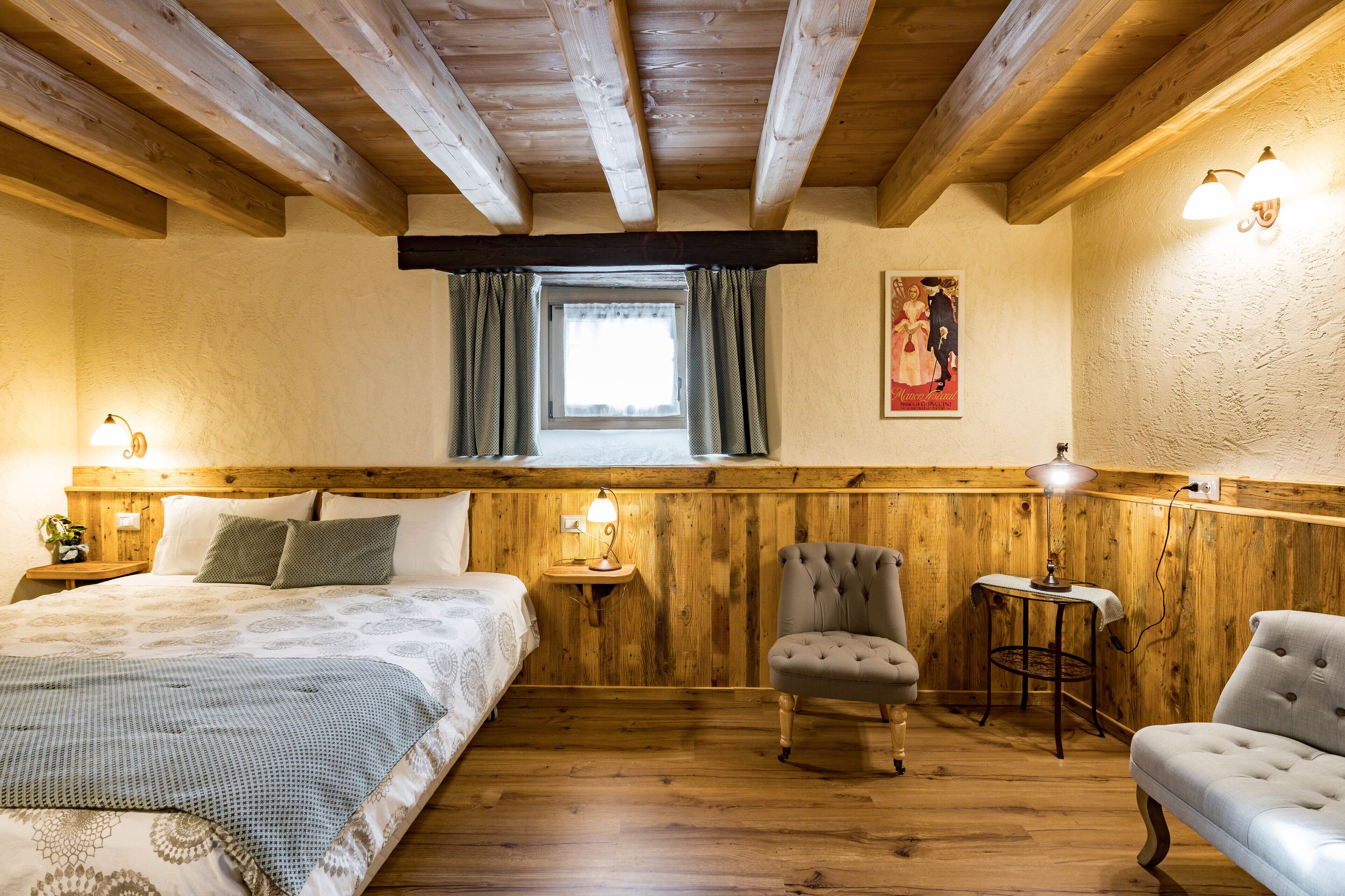 Een knus ingerichte kamer met houten plafond, een bed met beddengoed met patroon, een stoel en een kleine tafel met een lamp.