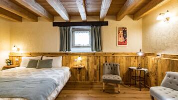 Een knus ingerichte kamer met houten plafond, een bed met beddengoed met patroon, een stoel en een kleine tafel met een lamp.