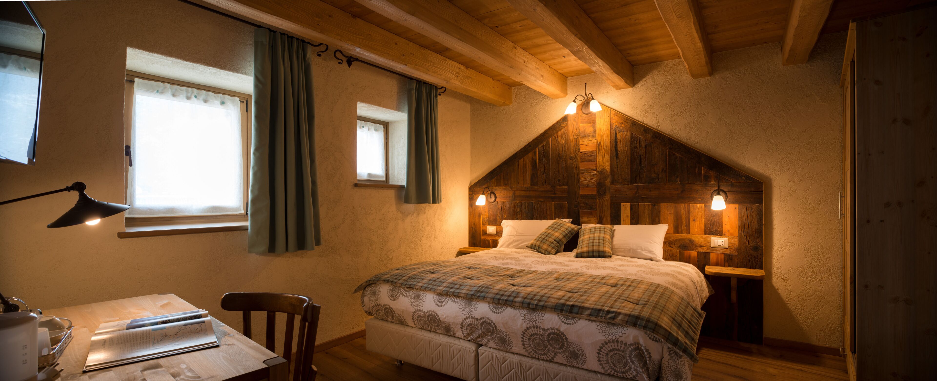 Un dormitorio acogedor con techo inclinado, cabecera de madera, cama con colcha escocesa, escritorio con lámpara y ventana con cortinas.