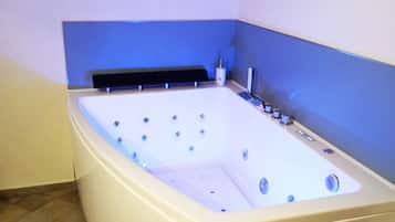Una bañera grande y rectangular con múltiples jets, un cabezal de ducha de mano y un panel de control. El cuarto tiene piso de baldosas y una ventana con cortina.
