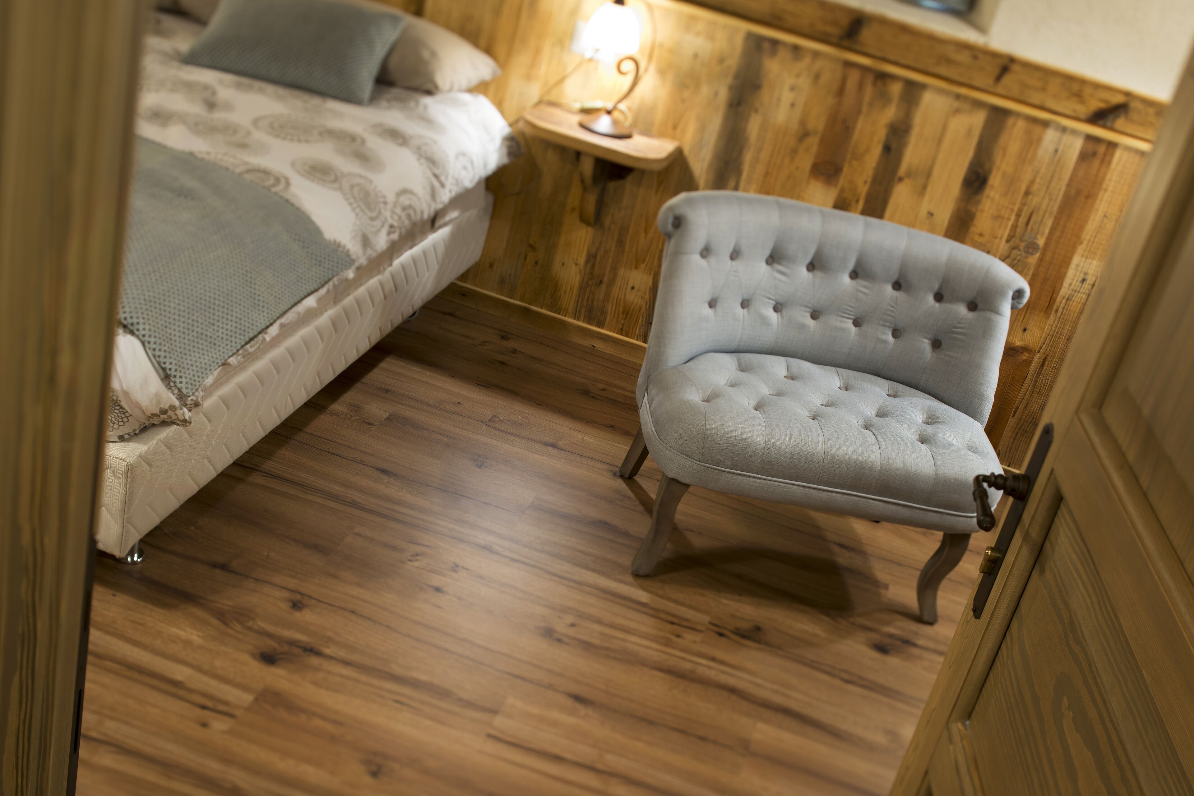 A room with a bed, a small table with a lamp, and a tufted armchair.