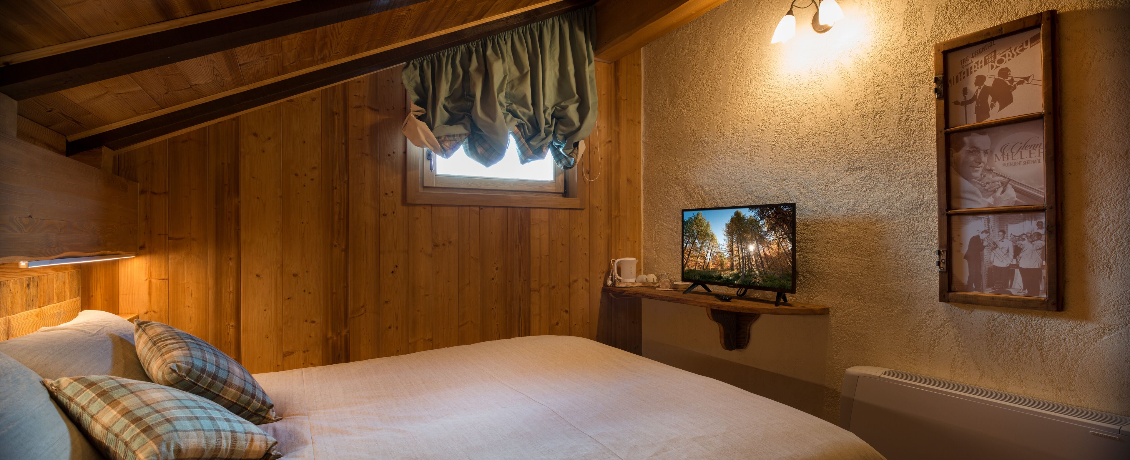 A cozy room with wooden walls, a bed with plaid pillows, a television on a wooden shelf, and a window with a curtain.