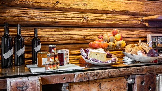 Una pared de madera rústica con un mostrador de madera que exhibe botellas, frascos, queso, frutas y pan.