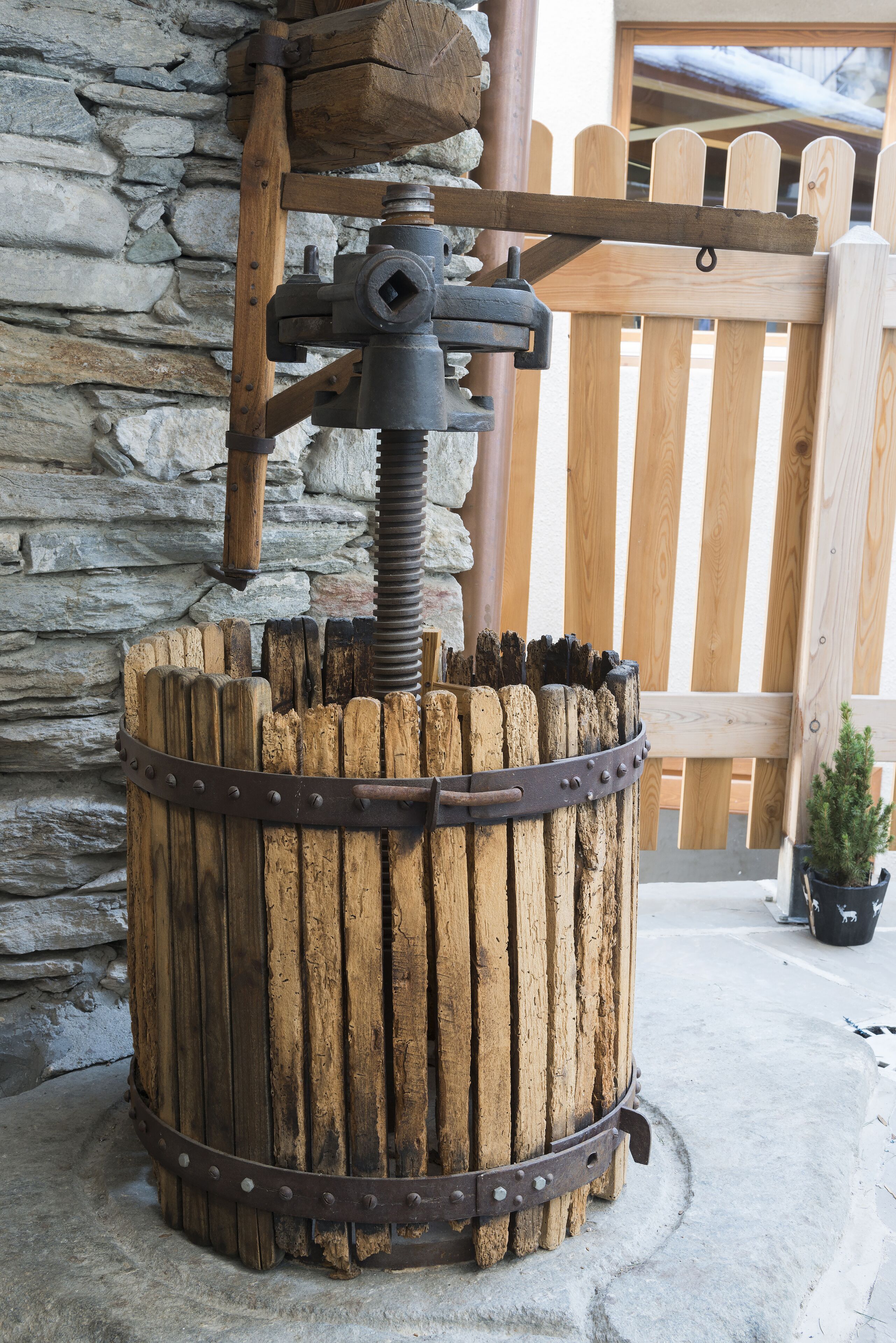 A rustic wooden barrel with a metal mechanism, set against a stone wall and wooden fence.
