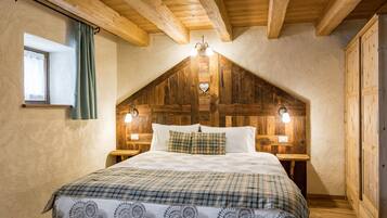 Une chambre mansardée chaleureuse, avec un plafond en pente en bois, un lit recouvert d’une couette à carreaux, deux lampes de chevet et une fenêtre avec des rideaux.