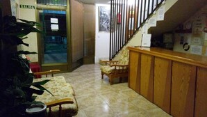 A reception area with a wooden counter, two upholstered chairs, a small table, and a staircase with a metal railing. - Hostal Mari Loli (San Fulgencio)