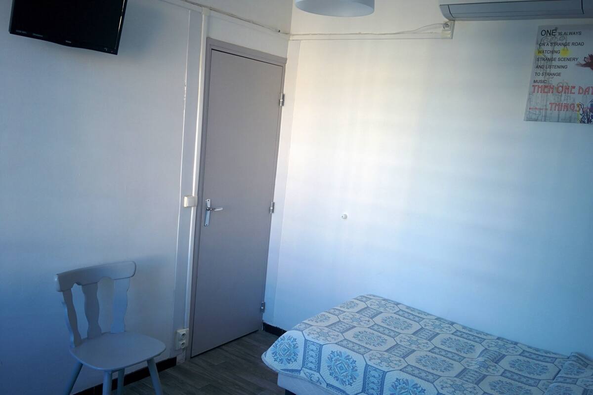 A small, sparsely furnished room with a single bed, a chair, and a door.
