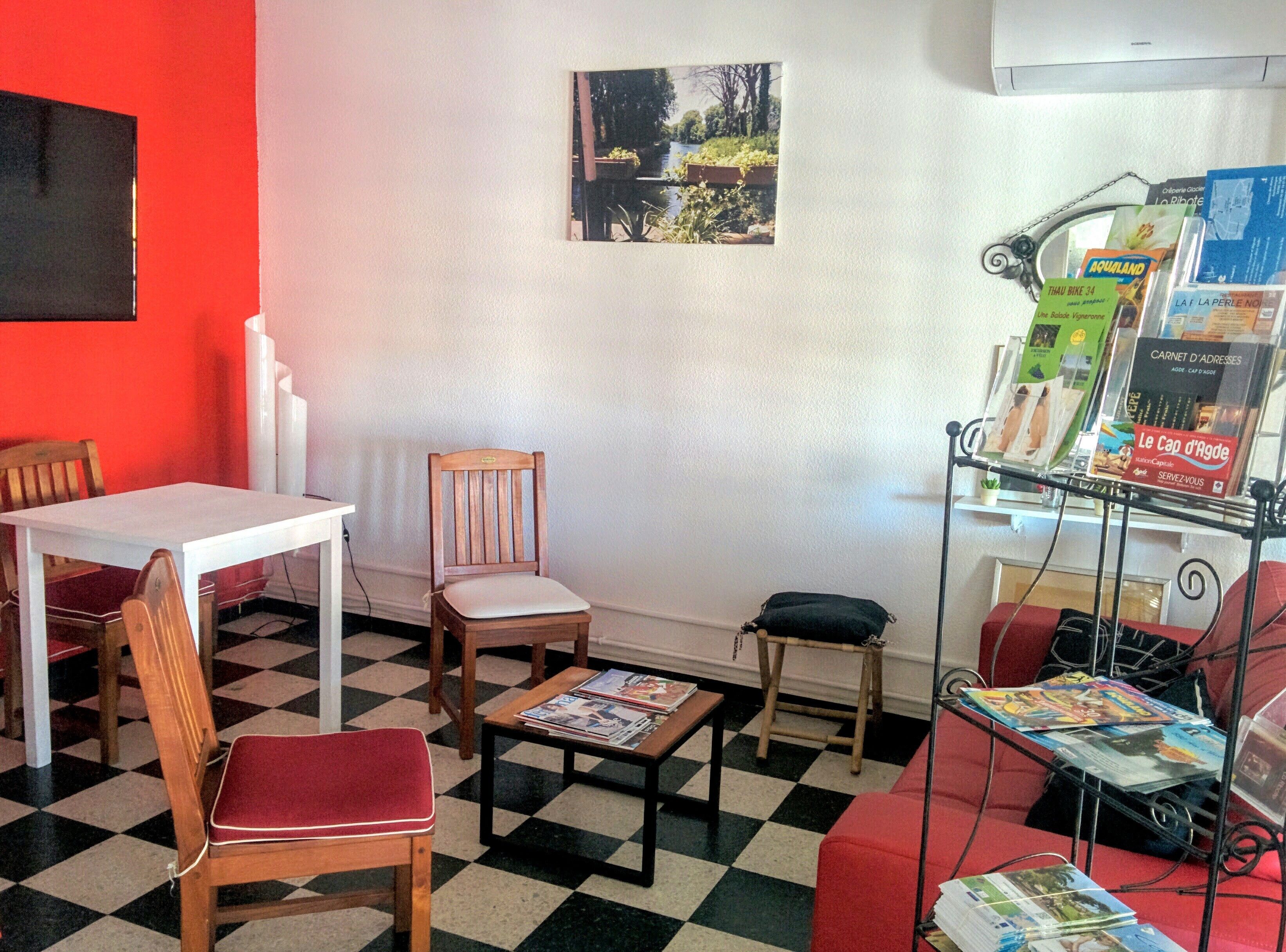 A room with a checkered floor, a white table, a wooden chair, a red sofa, and a magazine rack with various publications.