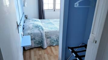 Una habitación pequeña y funcional con cama, una mesita, cortina de ducha y ventana con cortinas.