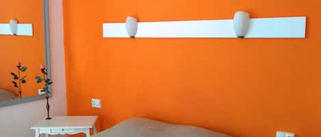 Un dormitorio con pared naranja, una mesita de noche blanca, una cama con colchón gris y un tocador blanco.