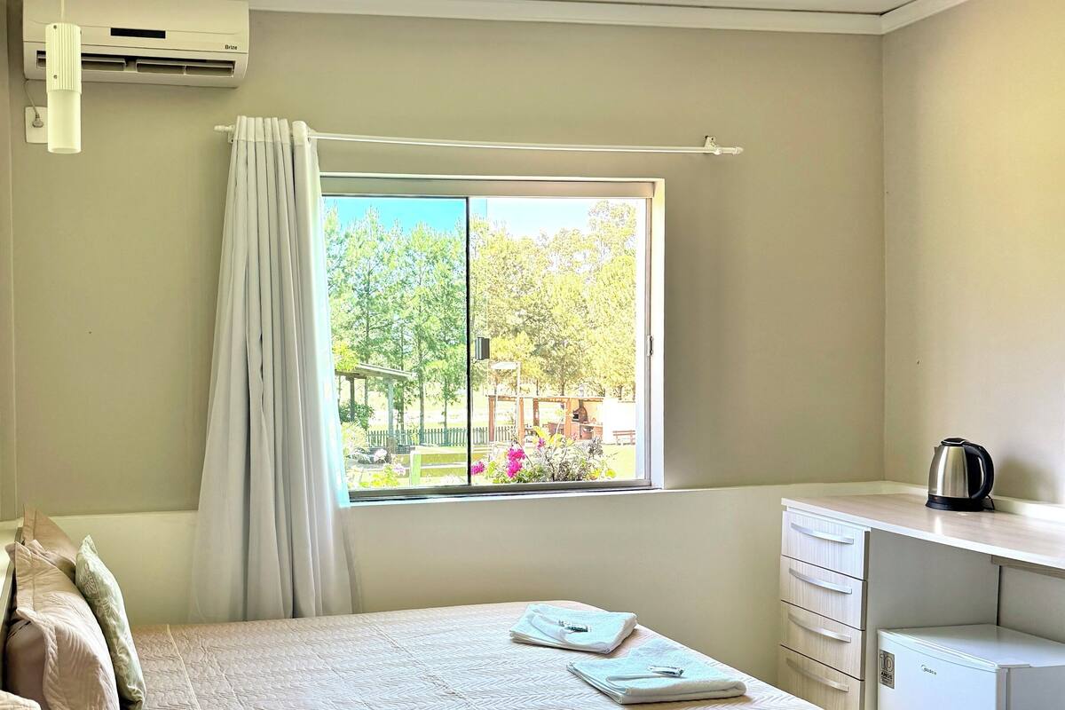 Une chambre d’hôtel avec deux lits, un bureau, un radiateur et une fenêtre donnant sur des arbres et un parc.
