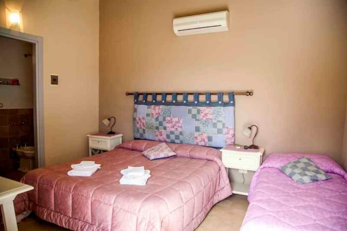 A bedroom with two beds, a headboard with a floral pattern, bedside tables with lamps, and an air conditioning unit on the wall.