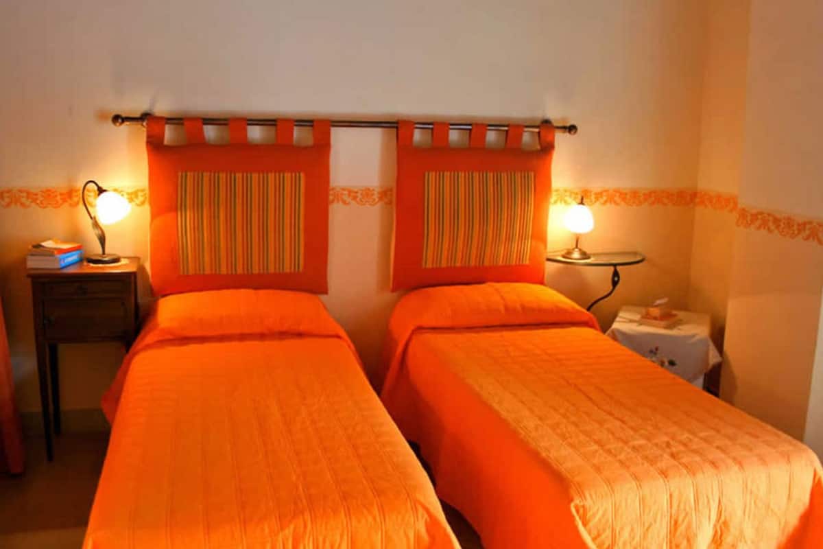 A room with two beds, each with an orange bedspread, a bedside table with a lamp, and a small table with a lamp.