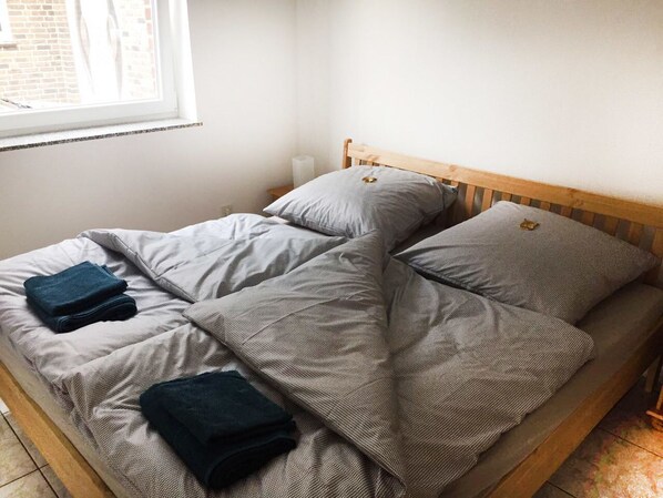 Un lit simple avec une tête de lit en bois, une literie grise et deux serviettes bleu foncé pliées.