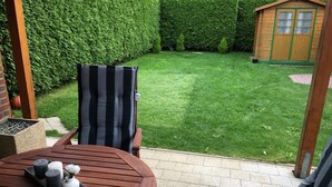 Un patio con una mesa redonda de madera, dos sillas a rayas y un pequeño jardín con seto y cobertizo de madera.