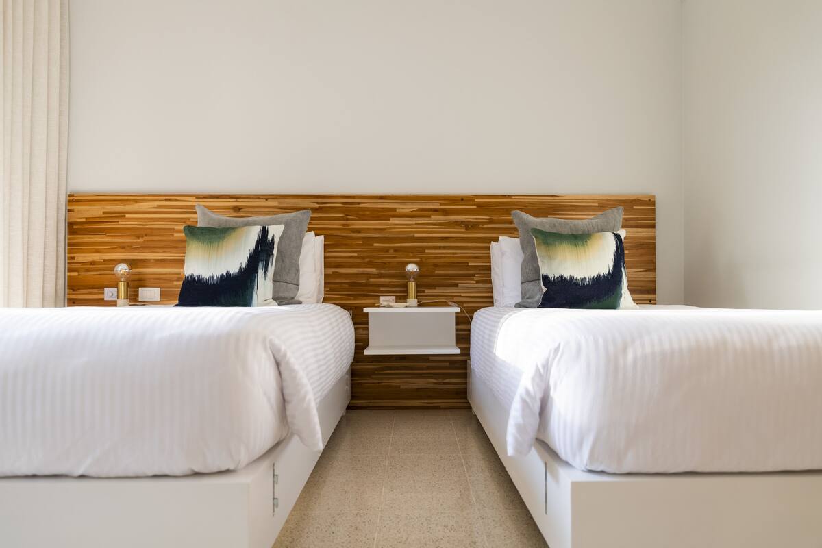 Quarto de hotel com duas camas, cabeceira de madeira, mesa de cabeceira pequena e prateleira fixada na parede.